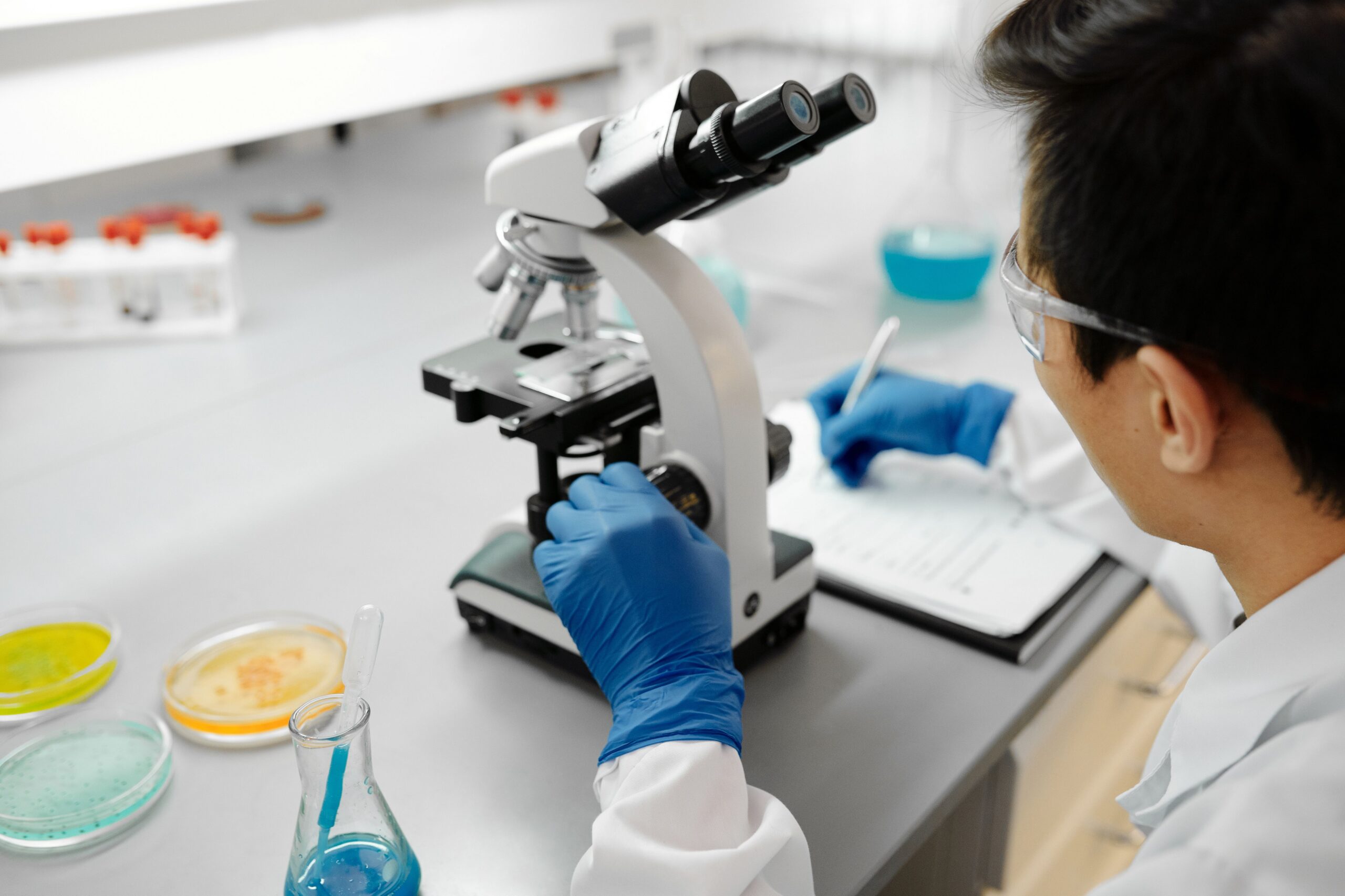 A laboratory analyst examining samples at a microscope, with petri dishes and a notebook on the bench.