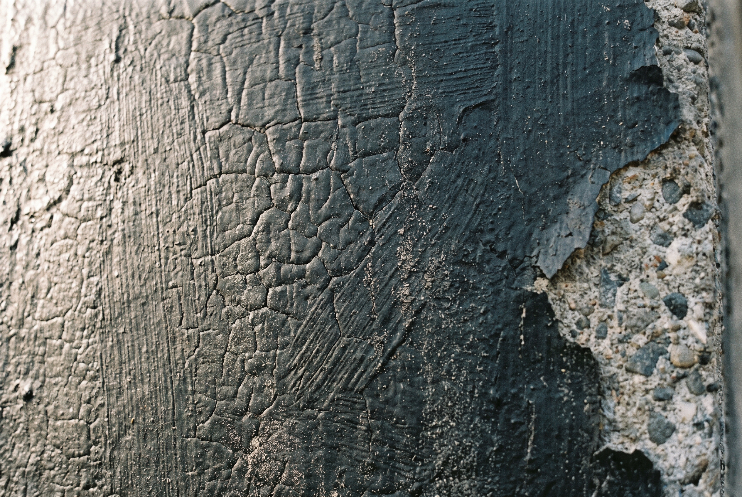 Close-up of an aged black tar coating on concrete, cracked into irregular plates and breaking away at one edge to expose the aggregate beneath.