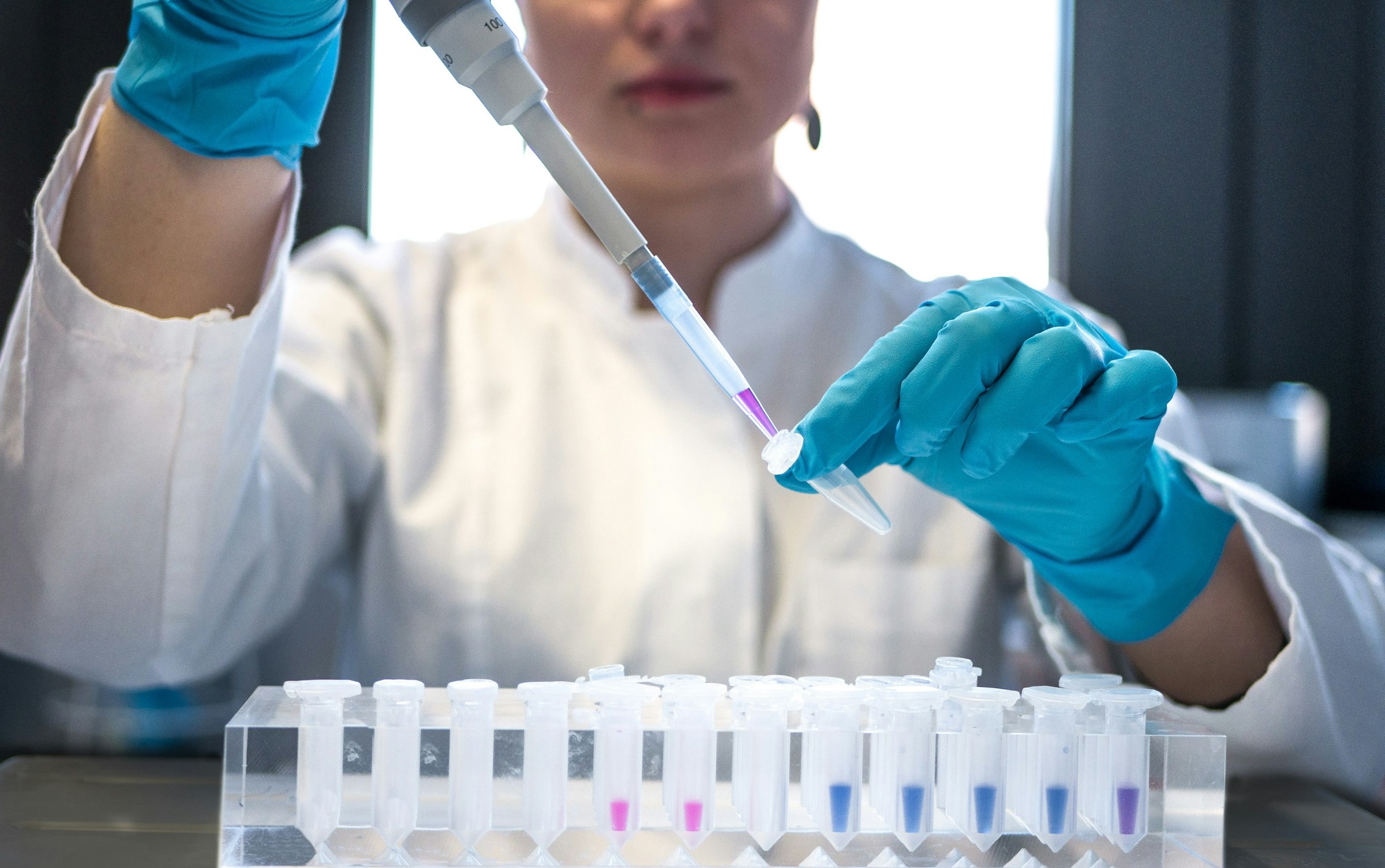 A laboratory technician in a white coat and gloves pipetting a sample into a rack of microtubes.