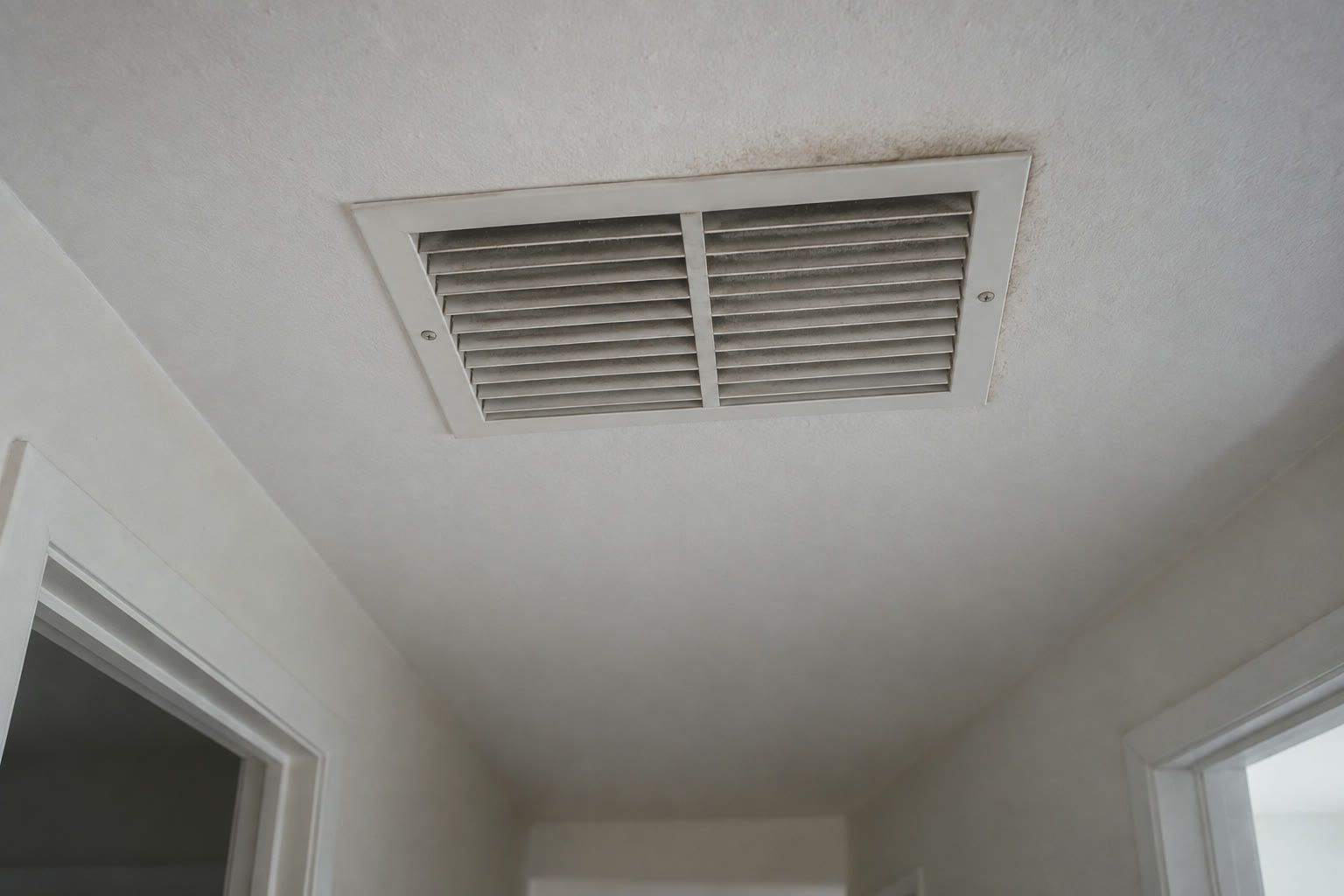 A ceiling air-supply grille with a film of dust on its louvers and faint discoloration on the paint around it.